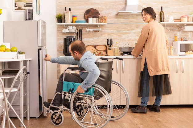 Mujer en la cocina mirando a su marido con discapacidad para caminar tratando de abrir la puerta del frigorífico. Hombre discapacitado paralítico discapacitado con discapacidad para caminar que se integra después de un accidente.