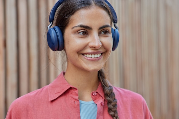 mujer con coleta tiene dientes blancos escucha la pista de audio en auriculares inalámbricos usa poses de camisa rosa