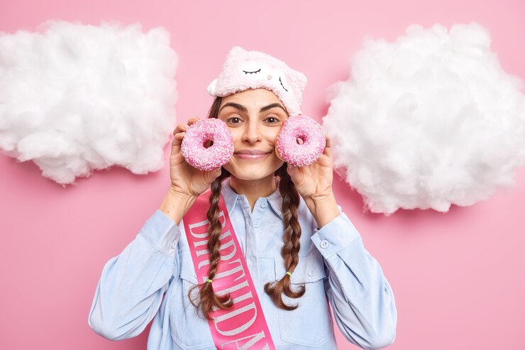 Foto gratuito mujer con coletas sostiene donas glaseadas cerca de la cara quiere comer productos de panadería usar antifaz en la frente camisa y cinta palabra escrita poses de cumpleaños alrededor de nubes blancas