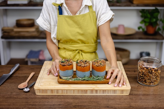 Mujer con coloridos postres dulces saludables budines de chía hechos de leche de almendras, extracto de espirulina azul, semillas de chía, mermelada de mango pappaya y granola casera. En la mesa de madera en la cocina de casa.