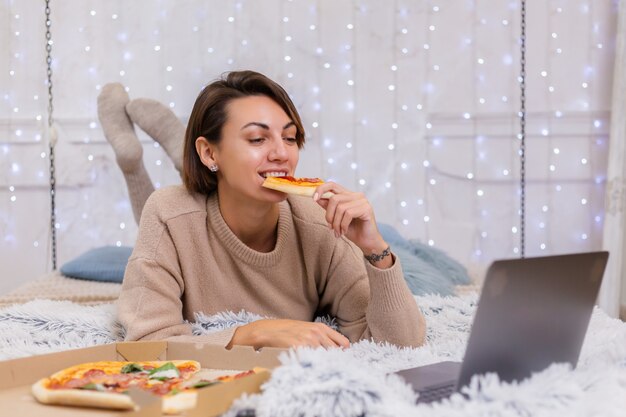 Mujer de comida rápida al este de la entrega en la cama en el dormitorio en casa. Mujer sola disfrutando de comida grasa, pizza