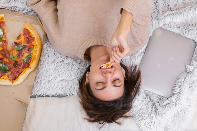 Mujer de comida rápida al este de la entrega en la cama en el dormitorio en casa. Mujer sola disfrutando de comida grasa, pizza