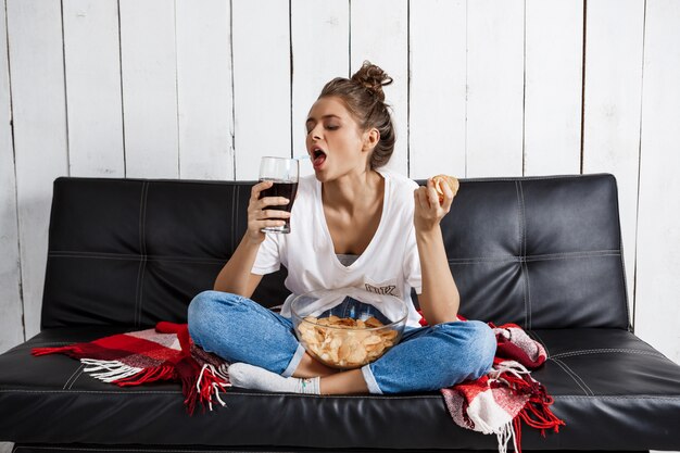 Mujer comiendo patatas fritas, bebiendo refrescos, viendo televisión, sentado en el sofá.
