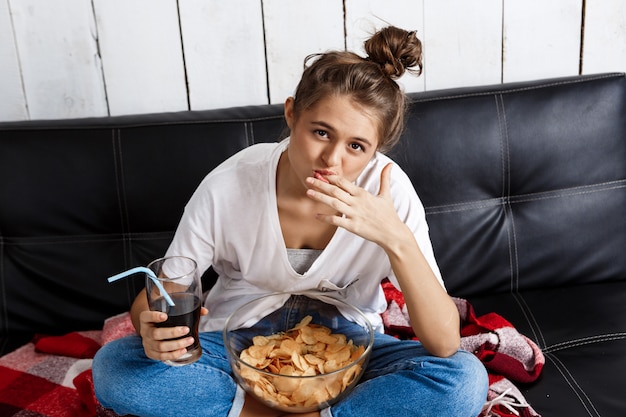 Mujer comiendo patatas fritas, bebiendo refrescos, viendo televisión, sentado en el sofá.