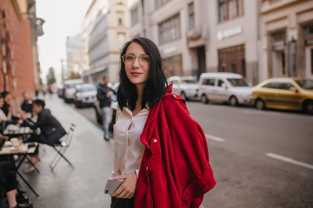 Mujer complacida con gafas de pie en la calle con teléfono en mano