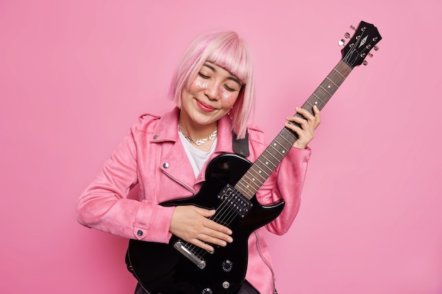 Mujer complacida de pelo rosa inclina la cabeza toca la guitarra acústica disfruta de la melodía favorita se prepara para el concierto de rock n roll viste poses de ropa de moda en el estudio contra la pared rosada. Concepto de musica