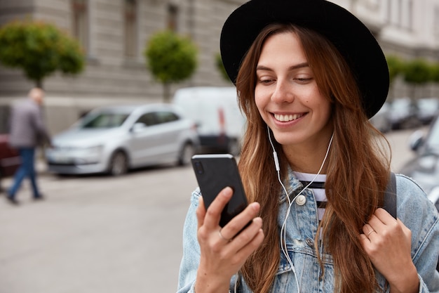 Mujer complacida con sombrero, sostiene el teléfono celular en la mano, escucha música de la lista de reproducción, pasea durante el tiempo libre por el entorno urbano