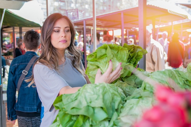 Mujer de compra de frutas y hortalizas en el mercado local de alimentos. Puesto de mercado con variedad de vegetales orgánicos. Retrato de mujer joven hermosa que elige verduras de hoja verde