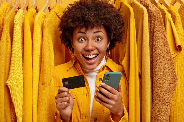 Mujer de compras alegre y emotiva utiliza el teléfono móvil para pagar en línea, tiene tarjeta de crédito, se encuentra entre suéteres amarillos en perchas
