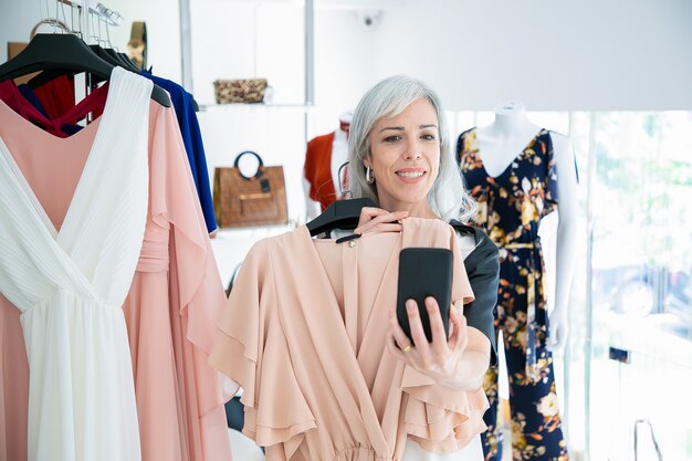 Mujer de compras en una tienda de moda y consultor amigo por teléfono móvil, mostrando el vestido elegido. Tiro medio. Concepto de comunicación o cliente boutique