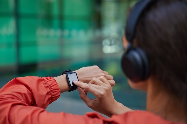 Mujer comprueba los resultados del entrenamiento físico en SmartWatch escucha música a través de auriculares vestidos con anorak plantea en borrosa