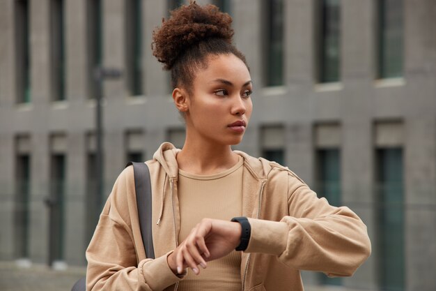 Mujer comprueba el tiempo en el reloj inteligente digital rastrea su desempeño wotkout mira hacia otro lado vestida con ropa deportiva cómoda posa contra el edificio de la ciudad