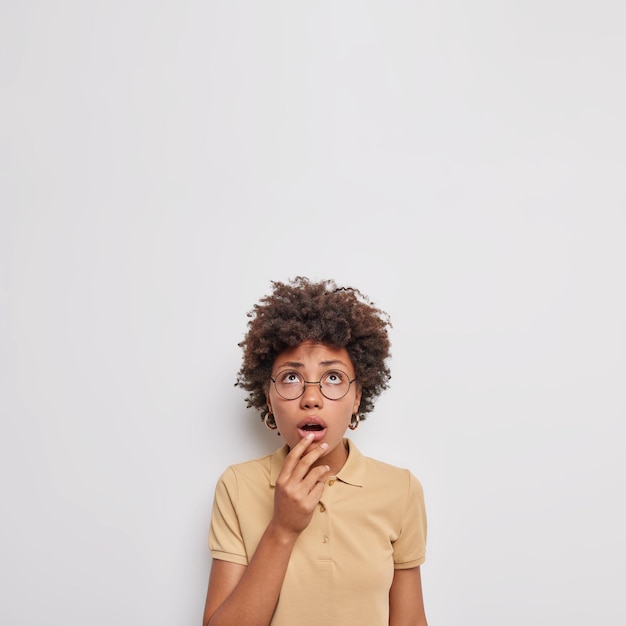 mujer concentrada arriba mantiene la boca abierta se siente asustada no puedo creer en algo impactante usa lentes transparentes camiseta casual marrón aislada en blanco. Oh no, que espantoso