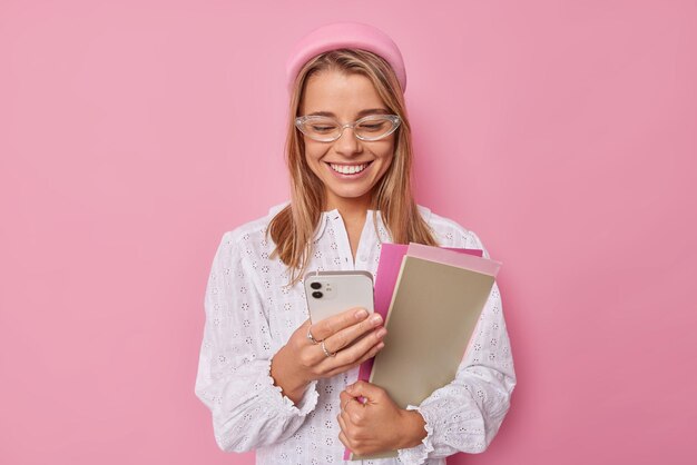 mujer concentrada con expresión alegre en la notificación de cheques de teléfonos inteligentes sostiene libros de texto y blocs de notas usa anteojos blusa blanca aislada en rosa. Estudiante de interior