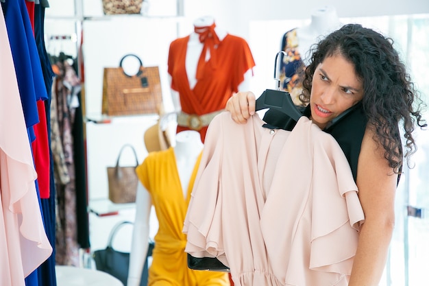 Mujer concentrada hablando por celular mientras compra en una tienda de moda, aplicando el vestido a sí misma y mirándose en el espejo. Tiro medio. Cliente boutique o concepto minorista