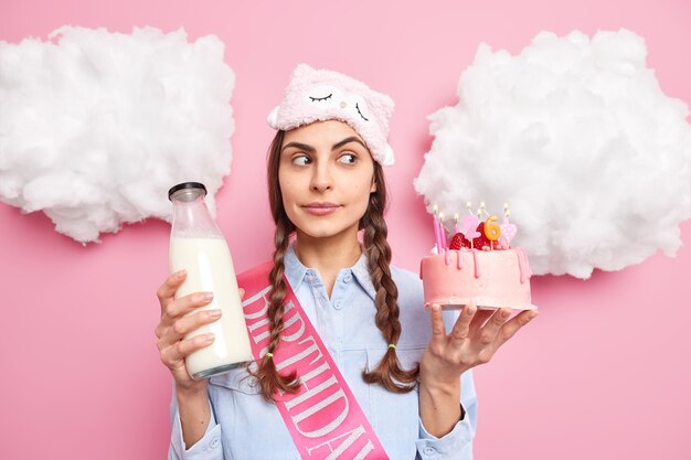 Mujer concentrada a un lado sostiene un delicioso pastel y una botella de leche que va a tener una cena festiva en su cumpleaños viste una camisa de dormir aislada en rosa