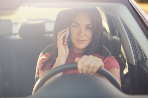 Mujer conduciendo un automóvil, tiene una conversación telefónica, se atasca en un embotellamiento, mira a través de winowshileld