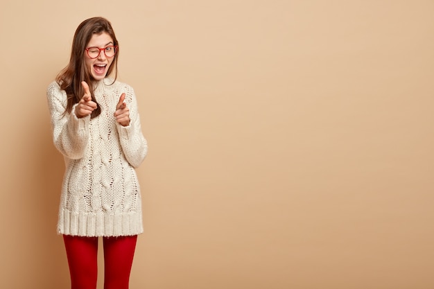 La mujer confiada y complacida parpadea, tiene una expresión facial segura, señala con ambos dedos índice, usa gafas, se para sobre una pared beige, copia espacio para anuncios o texto