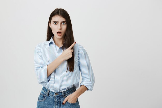 Mujer confundida en camisa y jeans apuntando hacia la esquina superior derecha