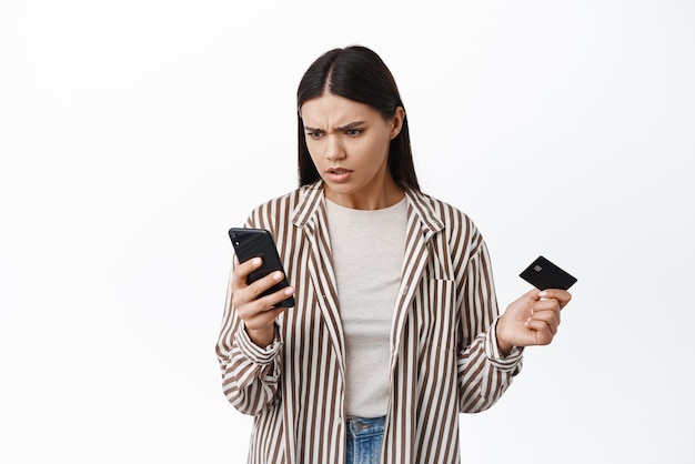 Una mujer confundida mirando su teléfono inteligente y sosteniendo una tarjeta de crédito de plástico no puede entender dónde está el dinero en la cuenta bancaria revisando el saldo o pagando el fondo blanco en línea