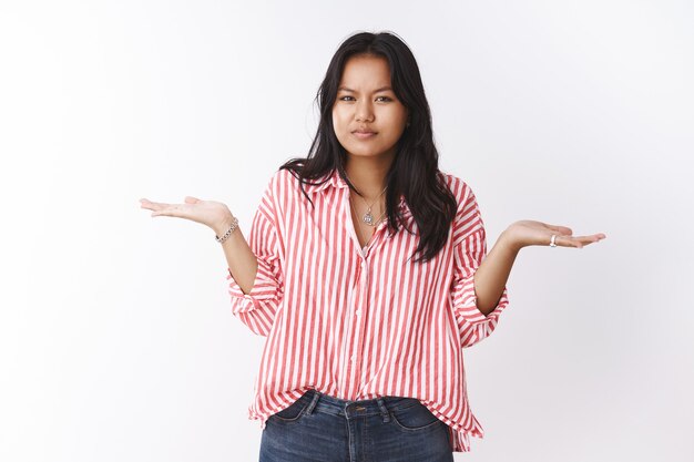 Mujer confundida de pie interrogada en pose despistada encogiéndose de hombros y levantando las manos en gesto incierto e inconsciente frunciendo el ceño, entrecerrando los ojos ante la cámara preocupada, no puede entender sobre la pared blanca