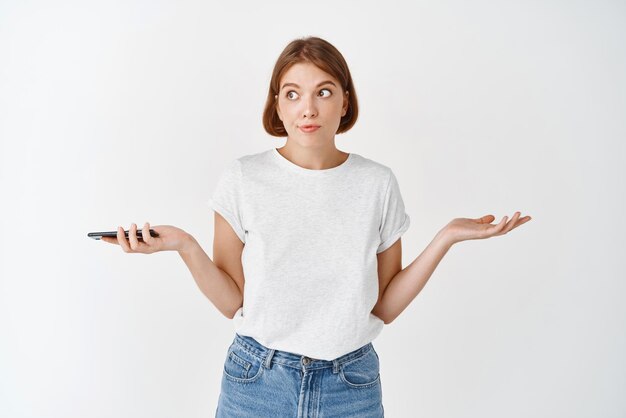 Mujer confundida sosteniendo el teléfono inteligente y encogiéndose de hombros sin darse cuenta de pie sin idea contra el fondo blanco