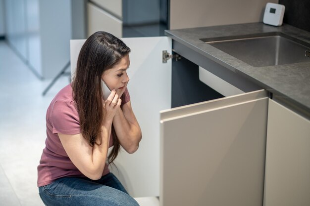 Mujer confundida con teléfono inteligente cerca de la oreja mirando debajo del fregadero