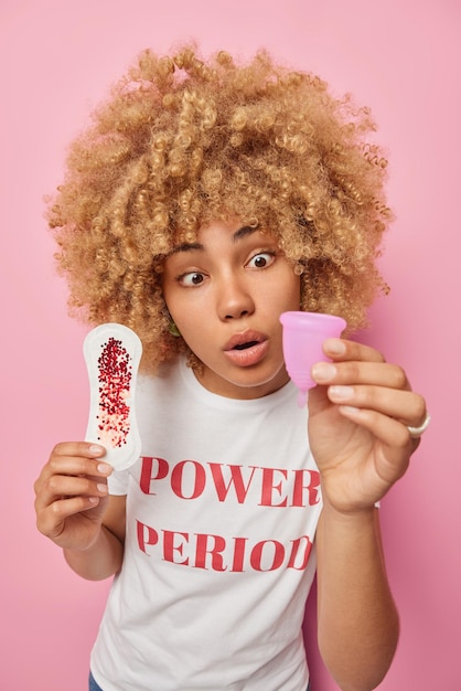 Una mujer conmocionada mira fijamente un producto de higiene desconocido que sostiene una taza de menstruación y una toalla sanitaria tiene días críticos vestida con una camiseta informal aislada sobre un fondo rosa Cuidado de la salud femenina