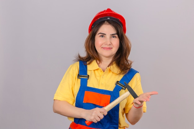 Mujer de constructor en construcción uniforme y casco de seguridad con sonrisa con martillo en manos de pie sobre la pared blanca aislada