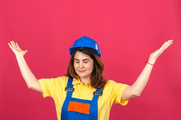 Mujer de constructor en uniforme de construcción y casco de seguridad de pie con los ojos cerrados encogiéndose de hombros extendiendo las manos sin entender lo que pasó sin expresión y confundida expresión sobre aislado