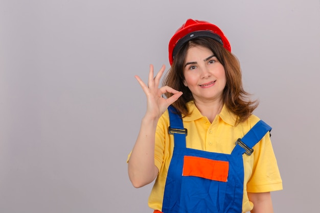 Mujer de constructor en uniforme de construcción y casco de seguridad con una sonrisa en la cara haciendo un signo bien parado sobre una pared blanca aislada con espacio de copia