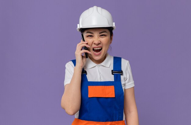 Mujer constructora asiática joven molesta con casco de seguridad blanco gritando a alguien en el teléfono