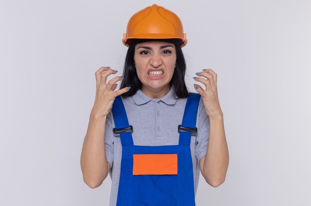 Mujer constructora joven enojada en uniforme de construcción y casco de seguridad gritando con los brazos levantados volviéndose salvaje