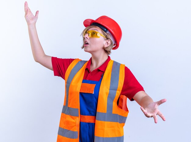 Mujer constructora joven sorprendida en uniforme con gafas extendiendo las manos aisladas sobre fondo blanco.