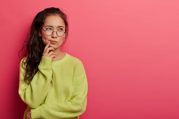 Mujer contemplativa mira seria y pensativa a un lado, frunce los labios y se concentra arriba, vestida con un jersey holgado verde, toma una decisión en mente