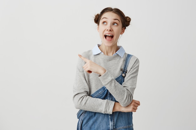 Mujer contenta en mono de jeans prestando atención a algo increíble que muestra el dedo índice de lado a lado con la boca abierta. Joven mujer encantada de presentar su lugar favorito. Copia espacio