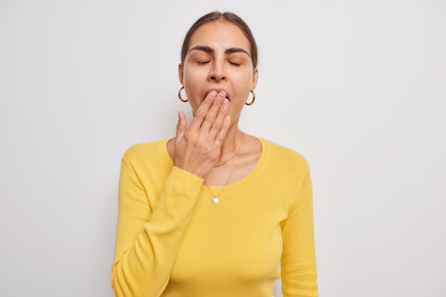 La mujer contra la boca con la mano mantiene los ojos cerrados viste un jersey amarillo casual siente fatiga o somnoliento se encuentra en blanco