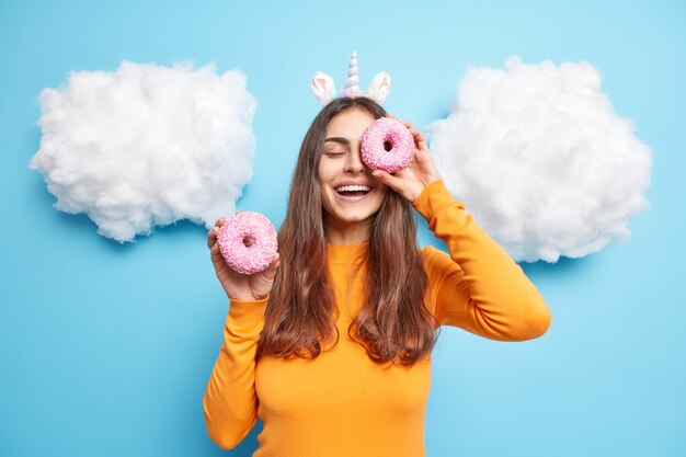 La mujer contra los ojos con deliciosas donas glaseadas sonríe positivamente tiene golosos viste un puente naranja aislado en azul