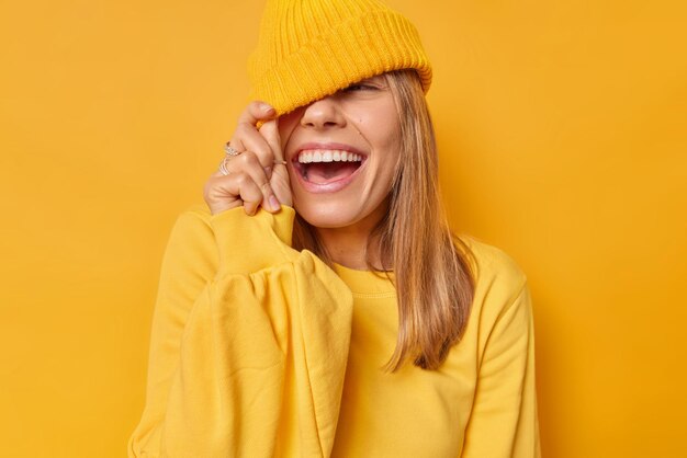 La mujer contra los ojos con sombrero tiene una expresión alegre vestida con un jersey casual aislado en amarillo brillante. Adolescente despreocupada tonta tiene expresión divertida