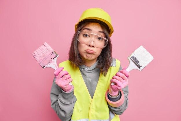 La mujer contratista sostiene dos pinceles para pintar en las manos, los labios, los labios, listos para pintar, las paredes interiores, usa casco de seguridad, lentes transparentes y uniforme