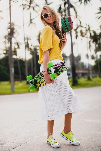 Mujer cool hipster con patineta y gorra posando sonriendo de vacaciones