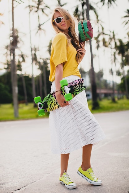 Mujer cool hipster con patineta y gorra posando sonriendo de vacaciones