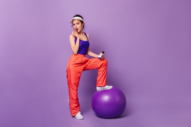 Mujer coqueta en traje deportivo posando en la pared púrpura con fitball