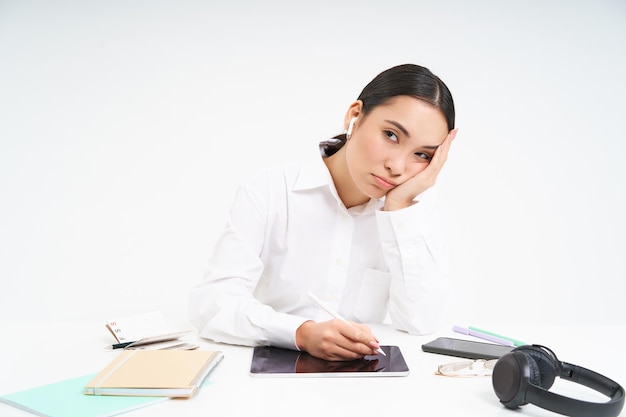 La mujer coreana cansada y aburrida en la oficina se sienta con una tableta digital y escucha música en los auriculares