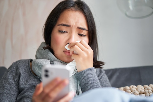 Mujer coreana enferma sentada en casa estornudando resfriada leyendo noticias en el teléfono móvil congelándose en su h