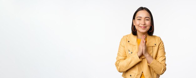 Mujer coreana feliz que parece esperanzada pidiendo ayuda a favor de la mendicidad de pie con gesto de namaste y sonriendo de pie sobre fondo blanco.