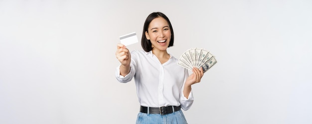 Mujer coreana feliz con tarjeta de crédito y dólares de dinero sonriendo y riendo posando contra el fondo blanco del estudio