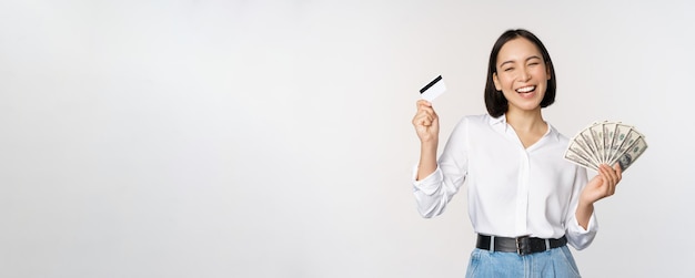 Mujer coreana feliz con tarjeta de crédito y dólares de dinero sonriendo y riendo posando contra el fondo blanco del estudio