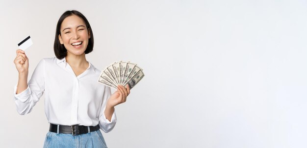 Mujer coreana feliz con tarjeta de crédito y dólares de dinero sonriendo y riendo posando contra el fondo blanco del estudio