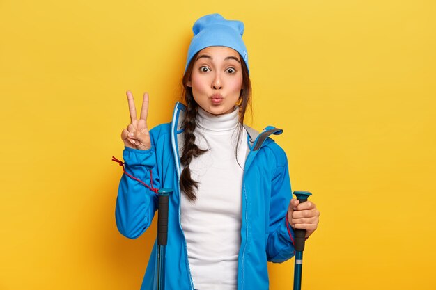 Mujer coreana morena hace un gesto de paz, caminatas en el bosque, sostiene bastones de trekking, vestida con ropa casual azul, disfruta de un descanso activo, posa sobre una pared amarilla